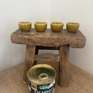 May include: Four small, olive-green ceramic bowls arranged in a row on a rustic, weathered wooden stool. In the foreground, a metal can of Maison Larzul Escargots de Bourgogne is visible, featuring a pull-tab lid and a black and gold label.