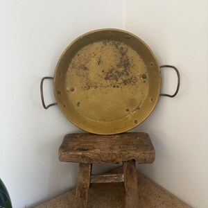 May include: A round, shallow brass pan with two curved metal handles on opposite sides. The interior surface of the pan shows a weathered, tarnished patina with dark spots and visible rivets securing the handles to the main body of the vessel.