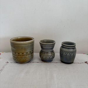 Puede incluir: Tres pequeños jarrones de cerámica con esmalte verde y azul. Los jarrones tienen diferentes diseños y están dispuestos en fila sobre un mantel blanco.