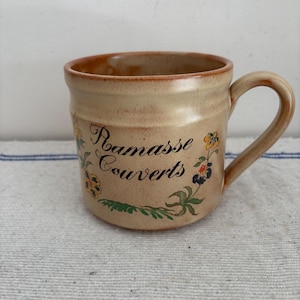 May include: Tan ceramic mug with a curved handle, featuring the text "Ramasse Couverts" and floral designs in yellow, blue, and green. The mug has a rustic, handmade appearance, with a slightly rough texture and a brown rim.