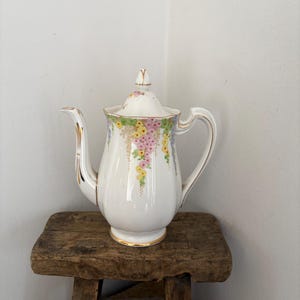 May include: A white ceramic coffee pot with a floral pattern in pink, yellow, and green. The pot has gold trim and a curved handle. It sits on a small, rustic wooden stool.
