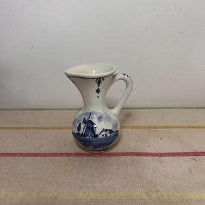 May include: A small, white ceramic jug with a blue and white Delft design. The jug features a windmill and a house in a landscape scene.