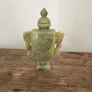 Puede incluir: Una pequeña urna antigua de color jade con tapa y dos asas ornamentadas. La urna presenta diseños florales tallados y se apoya sobre una base cuadrada. La tapa está rematada con un pináculo puntiagudo. La urna se exhibe sobre una superficie de madera envejecida.