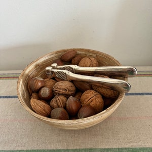 Peut inclure: Un bol en bois rempli de noix, dont des amandes, des noisettes et des noix. Un casse-noisette en argent repose sur les noix.