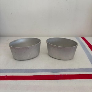 May include: Two small, oval-shaped, silver-colored metal containers. The containers are sitting on a white surface with red and blue stripes. The containers are empty and have a slightly tapered design.