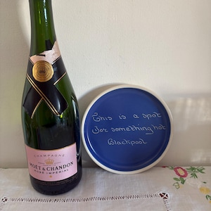 Op de afbeelding: Een blauwe keramische onderzetter met witte tekst die zegt "This is a spot for something hot Blackpool". Een fles Moët & Chandon Rosé Impérial champagne staat op een wit tafelkleed.