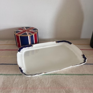 May include: A white rectangular serving tray with a blue and gold trim. The tray has a slightly scalloped edge. The tray is sitting on a striped tablecloth.
