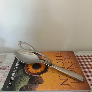 Puede incluir: Una cuchara de servir de plata con un mango largo apoyada en un libro de cocina titulado "The French Kitchen, A Cookbook" de Harris & Fran Warde.
