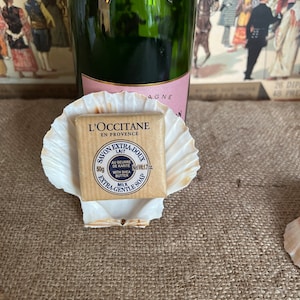 May include: A gold soap bar with the text "L'OCCITANE EN PROVENCE" and "SAVON EXTRA-DOUX LAIT" on the front. The soap is in a white seashell. The soap is 50g and is made with shea butter.