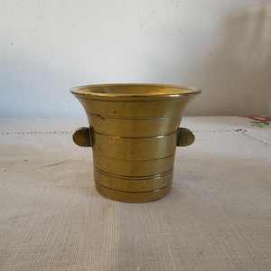 May include: A brass mortar with two small handles on the sides. The mortar has a smooth, shiny surface and is a light gold colour.