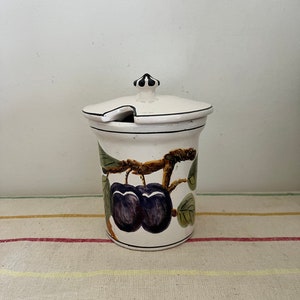 Op de afbeelding: Witte keramische pot met een deksel en een zwart-groen geverfd ontwerp van pruimen aan een tak.