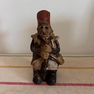May include: A small, hand-painted ceramic figurine of a soldier in a red hat and dark uniform. The figurine is holding a staff and has a detailed face. The figure is standing on a striped fabric.