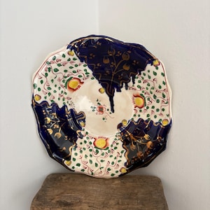 May include: A decorative ceramic plate with a scalloped edge. The plate features a cream-coloured centre with a floral pattern and is bordered by a deep blue design. The plate is resting on a rustic wooden stool.