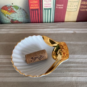 May include: A white seashell-shaped dish with a gold rim, sitting on a striped surface. A brown cork is in the center of the dish.
