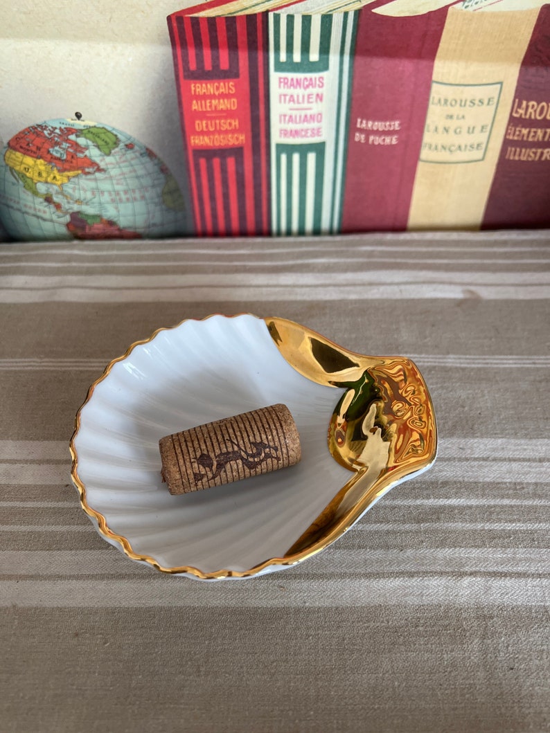 May include: A white seashell-shaped dish with a gold rim, sitting on a striped surface. A brown cork is in the center of the dish.