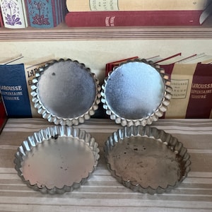 May include: Four silver tart pans with fluted edges. The pans are arranged in two rows of two on a striped beige surface.
