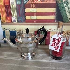 Może przedstawiać: Srebrny czajnik z czarnym uchwytem stoi na wzorzystej serwetce obok szklanej butelki wypełnionej czerwonymi zapałkami i białą etykietą z napisem "wesołych świąt".