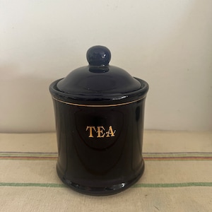 Könnte beinhalten: Eine schwarze Teedose aus Keramik mit goldenem Deckel und dem Wort "TEA" in goldenen Lettern.