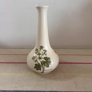 May include: A white ceramic vase with a floral design featuring green leaves and white flowers. The vase has a narrow neck and a rounded base. The text "Alchemilla vulgaris" is printed on the vase.