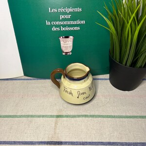 May include: A small, brown and white ceramic creamer with a brown handle. The creamer has the words "Fresh from the cow" written on it.