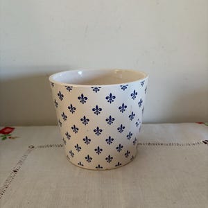 Może przedstawiać: Biała ceramiczna doniczka z powtarzającym się wzorem niebieskich symboli fleur-de-lis. Doniczka ma kształt cylindryczny i stoi na białym obrusie.