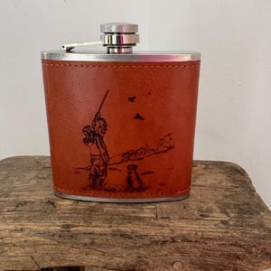 A vintage Selbrae House hip flask from stainless steel, hand-bound in brown leather with an engraved shooting-themed image of a man & dog.