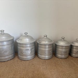 Pode incluir: Um conjunto de cinco latas de metal prateado de tamanhos graduados com tampas abobadadas a condizer. Cada recipiente cilíndrico apresenta detalhes horizontais canelados e uma placa de etiqueta retangular na parte frontal.