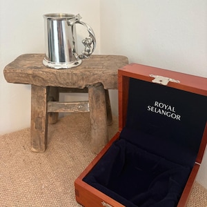 Peut inclure: Une chope en argent avec une poignée en forme d'ours en peluche est posée sur un tabouret en bois rustique. À côté, une boîte en bois ouverte avec le texte "ROYAL SELANGOR" à l'intérieur. La boîte a une doublure en velours bleu foncé.