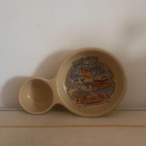May include: A beige ceramic dish with a side compartment. The dish features a colourful illustration of a seaside town with the text "Côte Vendéenne".