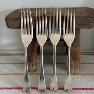 May include: Four silver forks with tines, arranged in a row. The forks have long, slender handles and a heart-shaped design at the base. They are placed on a striped tablecloth with a wooden surface in the background.