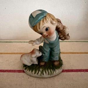 May include: A porcelain figurine of a boy wearing a blue cap and overalls, holding a brown object on his back, and looking down at a white puppy.