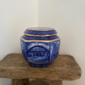 Könnte beinhalten: Achtkantige blau-weiße Keramikdose mit Deckel und Goldrand, verziert mit einer Brückenillustration. Die Dose, mit Text am Boden, steht auf einem rustikalen Holzschemel. Das Design ist klassisch.