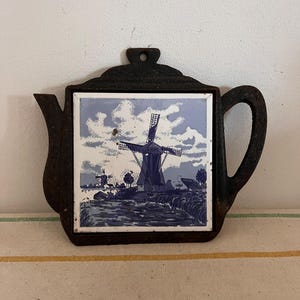 Puede incluir: Un posavasos de tetera de hierro fundido negro con una baldosa azul y blanca con un molino de viento y un cielo nublado.