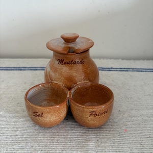 May include: A ceramic condiment set with a mustard pot and two attached bowls labeled "Sel" and "Poivre." The mustard pot has a lid and is labeled "Moutarde." The set is a warm brown color.