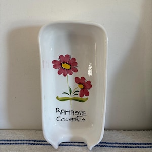 May include: White ceramic spoon rest with a floral design. The spoon rest features two red flowers with yellow centers and green stems. The words "RAMASSE COUVERTS" are written in black script. The spoon rest has a rectangular shape with a curved top and two small handles.