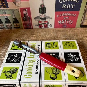 Może przedstawiać: Narzędzie kuchenne z czerwoną i białą rączką oraz srebrnym metalowym ostrzem. Narzędzie leży na książce kucharskiej zatytułowanej "Cooking Explained" autorstwa Barbary Hammond. Książka kucharska ma zielono-biały projekt z ilustracjami jedzenia.