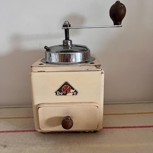 Op de afbeelding: Een vintage witte koffiemolen met een houten handvat en een metalen kruk. De molen heeft een rood en blauw windmolenlogo met de tekst "De Ve" aan de voorkant.