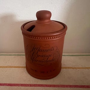 May include: A terracotta jar with a lid, featuring the text "St. James's Orange Marmalade" in a decorative script. The jar has a brown and white striped background.