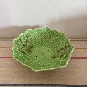 May include: A green ceramic colander shaped like a cabbage leaf with brown painted details. The colander has small holes throughout the bottom.