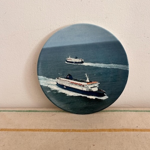 May include: A round ceramic plate with a blue and white image of two ferries sailing on the ocean.