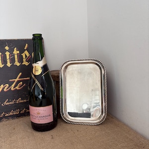 Puede incluir: Una bandeja rectangular de metal plateado con un borde trenzado tipo cuerda, colocada junto a una botella de vidrio verde oscuro de champán Moët & Chandon Rosé Impérial con etiqueta rosa y cuello de lámina dorada.