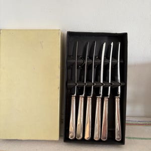 May include: A set of six silver-handled knives in a black box with a cream-colored lid. The knives have a simple, elegant design with a shiny, reflective finish. The box is rectangular and provides a neat storage solution.