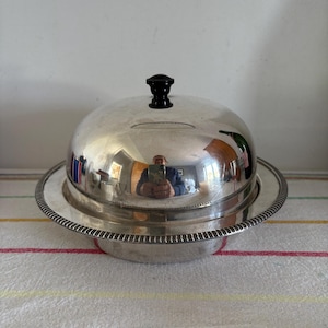 Könnte beinhalten: Eine silberfarbene Butterdose mit gewölbtem Deckel und schwarzem Griff. Die Schale steht auf einem runden Sockel mit dekorativem Perlenrand. Die Butterdose steht auf einer gestreiften Tischdecke.