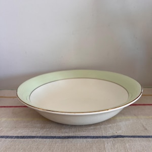 Może przedstawiać: Biała ceramiczna miska z zielonym i złotym obrzeżem. Miska stoi na wzorzystej serwetce.