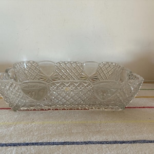 May include: A clear, rectangular glass dish with a diamond-cut pattern. The dish has a scalloped edge and heart-shaped designs on the sides. It sits on a striped fabric.