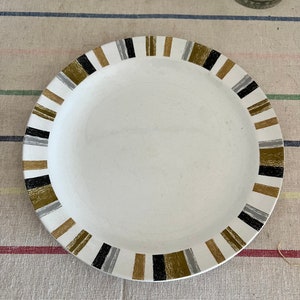 Peut inclure: Une assiette en c&eacute;ramique blanche avec une bordure ray&eacute;e noire, dor&eacute;e et grise. L'assiette est pos&eacute;e sur une surface en tissu ray&eacute;e.