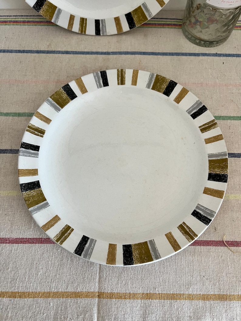 Peut inclure: Une assiette en c&eacute;ramique blanche avec une bordure ray&eacute;e noire, dor&eacute;e et grise. L'assiette est pos&eacute;e sur une surface en tissu ray&eacute;e.