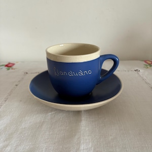 Può includere: Un set di tazza e piattino in ceramica blu. La tazza ha un bordo bianco e un manico. La parola "Llandudno" è scritta sulla tazza. Il piattino è anch'esso blu con un bordo bianco. Il set è su una tovaglia bianca.