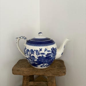 Peut inclure: Une théière en céramique bleue et blanche avec un couvercle, une anse et un bec verseur. La théière présente un motif Willow traditionnel. La théière est posée sur un petit tabouret en bois rustique.