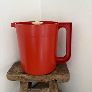 Könnte beinhalten: Ein roter Kunststoffkrug mit Griff und weißem Deckel. Der Krug steht auf einem kleinen, verwitterten Holzschemel. Der Krug hat einen Ausguss zum Ausgießen und ist von leuchtend roter Farbe.
